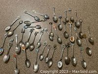 Overview photo showing the full collection of 37 souvenir spoons spread out on a brown surface, showing different sizes, handle designs and colorful emblems.