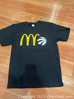 Full view of black T-shirt with McDonald's golden arches and Toronto Raptors claw basketball logos on front, laid flat on wooden floor.