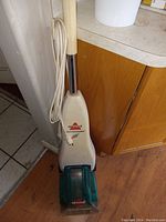 Front view of Bissell Quicksteamer carpet cleaner leaning against a wall, showing beige body with green base and power cord wrapped around handle.