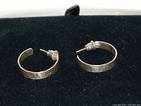 Pair of hand-hammered sterling silver hoop earrings shown on black and satin fabric backgrounds.