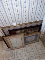 View of five framed artwork pieces stacked on the floor against a wall with floral wallpaper, showing varying frame sizes and styles.