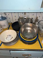 Photo showing a variety of metal bakeware including round cake pans, a large metal mixing bowl, a cast iron skillet, and a white decorative Pyrex serving bowl on a kitchen counter.