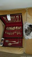 Wooden silverware box open displaying forks, spoons, knives, and serving pieces with a plastic bag of additional silverware items beside it on a couch.