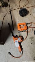 Photo showing the Black and Decker orange jig saw, the hedge trimmer blade, and the drain snake reel on a floor.