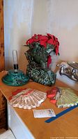 Photo showing green ceramic lamp base, two ceramic Native American face wall plaques, and green planter with red faux floral arrangement on wooden table