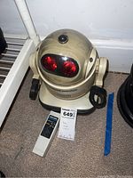 Front and angled view of Robie Junior robot showing red illuminated eyes and attached base, placed next to the remote control unit on the floor.