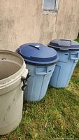 Three Rubbermaid Roughneck trashcans outdoors on grass near building wall, two with blue lids and one without lid visible