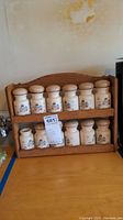 Photo of vintage ceramic spice jars with lids on wooden rack. One jar missing lid. Jars have painted scenes of houses and countryside.