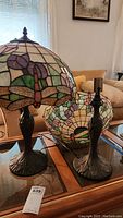 Pair of stained glass dragonfly lamps on a glass table, showing one lamp with complete shade and harp and the other lamp missing harp and with detached shade beside it.