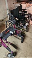 Photo showing maroon walker with black seat and hand brakes in front, with two black folding wheelchairs behind it in basement setting.