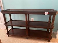 Front view of wooden console table showing three shelves, decorative columns, and caster wheels at the base.