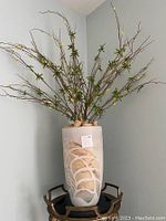 Artificial plant with thin branches and small green leaves in beige ceramic pot with decorative accents, photographed against a corner wall
