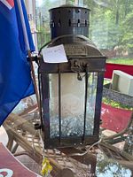 Photo of black metal decorative lantern containing a white floral candle resting on clear decorative pebbles inside a glass enclosure, placed on a glass table.