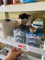 Shelf with three clear plastic stacked bins with blue lids filled with various metal hardware components, white plastic basket, cloth bag, and tissue boxes