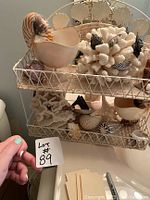 Photo of the full two-tiered wire shelf holding various nautical themed bathroom decor items including seashell shaped soaps, coral and shell decor pieces, small shell-framed mirror, tissue holder and trash bin