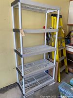 Full view of the 5-tier plastic storage shelf against a yellow wall, showing white slotted shelves and vertical poles with black connectors.