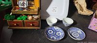 Two Japanese saki cups with their saucers placed on a dark surface beside a wooden jewelry box