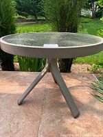 Small round outdoor table with textured glass top and light brown metal tripod base on tiled patio with garden plants in background.