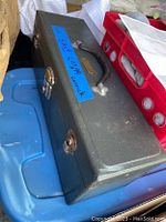 Closed dark gray metal toolbox with handle and blue tape label, showing external condition.