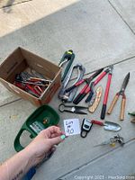 Photo showing assorted yard tools, hand pruners, metal shears, red handled bolt cutters or loppers, green hose spray nozzle, small garden scoop, boxed miscellaneous metal hand tools and sprinkler accessories on pavement.
