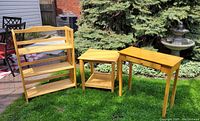 Three light-colored solid wood furniture pieces on grass: folding 3-shelf shelving unit, square end table with lower shelf, and rectangular table with drawer.