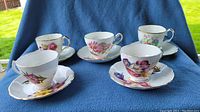 Five porcelain teacups each paired with matching saucers featuring different colorful floral patterns on a blue cloth background.