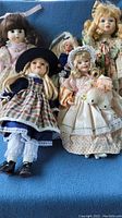 Five dolls arranged on blue fabric surface showing various dolls including Mary with lamb, Anne of Green Gables, Barbie, blonde and brunette dolls