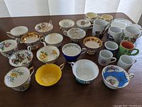 Front view of assortment of 25 cups and mugs, showing bone china and ceramic items with floral, gold, and graphic designs.