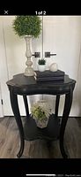 Full view of the decorative side table with dark painted finish. On top are decorative items including a tall candle holder with a green plant, two stacked books, a small white pot with a succulent, and a white decorative object. The lower shelf holds a glass vase with artificial flowers.