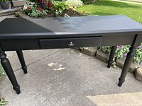 Rectangular dark finished painted table with one drawer and turned legs, shown outdoors on concrete with greenery in background.