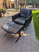 Black leather occasional chair with padded seat and back cushions and matching footstool, placed outdoors on a brick patio, showing overall condition and style.