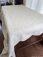 Full view of the cream colored quilt/bed cover displayed on a wooden surface showing scalloped edges and overall condition.