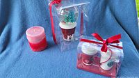 Photo of Starbucks Christmas snow globe, two coffee cup Christmas tree ornaments in clear packaging with red bows, and a red candle all displayed on a blue background.