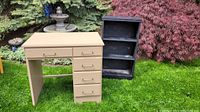 Photo of beige painted wooden desk and black painted wooden bookcase outdoors.