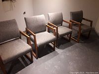 Four grey fabric office waiting chairs with wooden armrests arranged in a row on a concrete floor with a plain background.