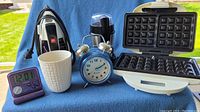 Wide-angle view of all items: Salton waffle maker open showing grids, black Proctor Silex iron, black and silver Black & Decker grinder behind, blue windup alarm clock in center, white handleless cup to the left of clock, small purple travel alarm clock and purple digital timer in front at left.