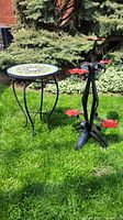 Side view of iron plant stand and mosaic topped table positioned on grass with greenery in background