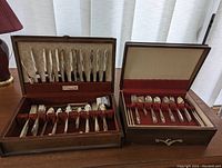 Two wooden cases open showing a large quantity of silverplate flatware organized neatly inside: one case holds 12 knives on the lid and various forks and spoons in the bottom, the other case holds grouped utensils including knives, forks, spoons, and extra pieces.