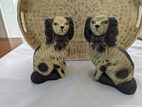 Pair of ceramic dog figurines on a table with a textured tray behind them, showing full side-front view of both dogs.