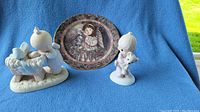 Front view of the two Precious Moments figurines and the collector plate on blue fabric.