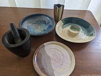 Overview of the full set: heavy dark stoneware mortar and pestle, bluish bowl, green and cream serving dish with central bowl, smaller brownish/cream platter, and small striped vase all on a wood table.