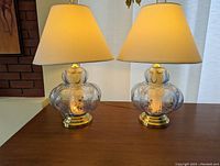 Two pale blue glass lamps with beige shades, sitting side by side on a wooden surface, both turned on showing diffused light.