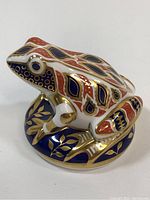 Side view of the porcelain frog paperweight showing intricate Imari pattern in cobalt, red, and gold accents on white porcelain base with frog sitting on decorated round base.