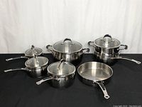 Full view of six Lagostina stainless steel pots and pans placed on a black surface, with four of them having glass lids and two without lids.