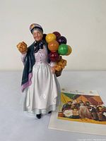 Front view of Royal Doulton 'Biddy Penny Farthing' figurine with a booklet beside it.