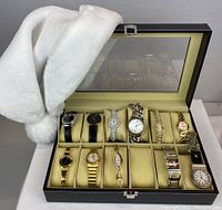 Twelve watches displayed in open black faux leather case with glass lid, white cloth draped