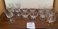 Full view of 14 etched glasses arranged on a wooden surface, showing different glass shapes including goblet and coupe styles.