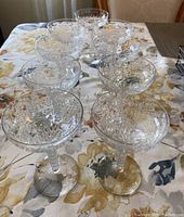 Nine antique etched crystal champagne saucer glasses arranged on a floral tablecloth showcasing the overall set and etching pattern.