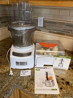 Full view of Cuisinart food processor with clear bowl, white base, control panel with buttons, and user manuals on the counter next to it.