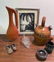 Photo showing all Mediterranean items arranged on wooden surface including MCM clay pitcher, leather tooled pitcher, grape bottle, stone barrel, ashtray, brass slippers, and lithograph.
